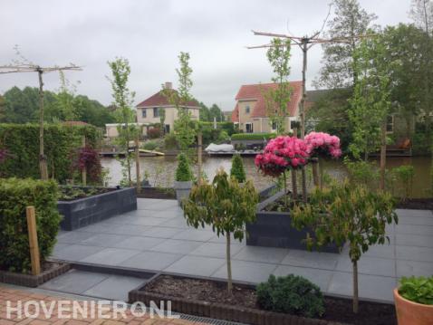 Strakke tuin aan het water met siertegels, plantenbakken en leibomen