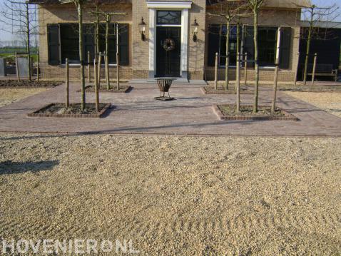 Villatuin met grind, bestrating van klinkers en jonge bomen.