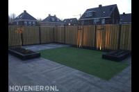 Achtertuin met bestrating, kunstgras, leibomen en houten schutting